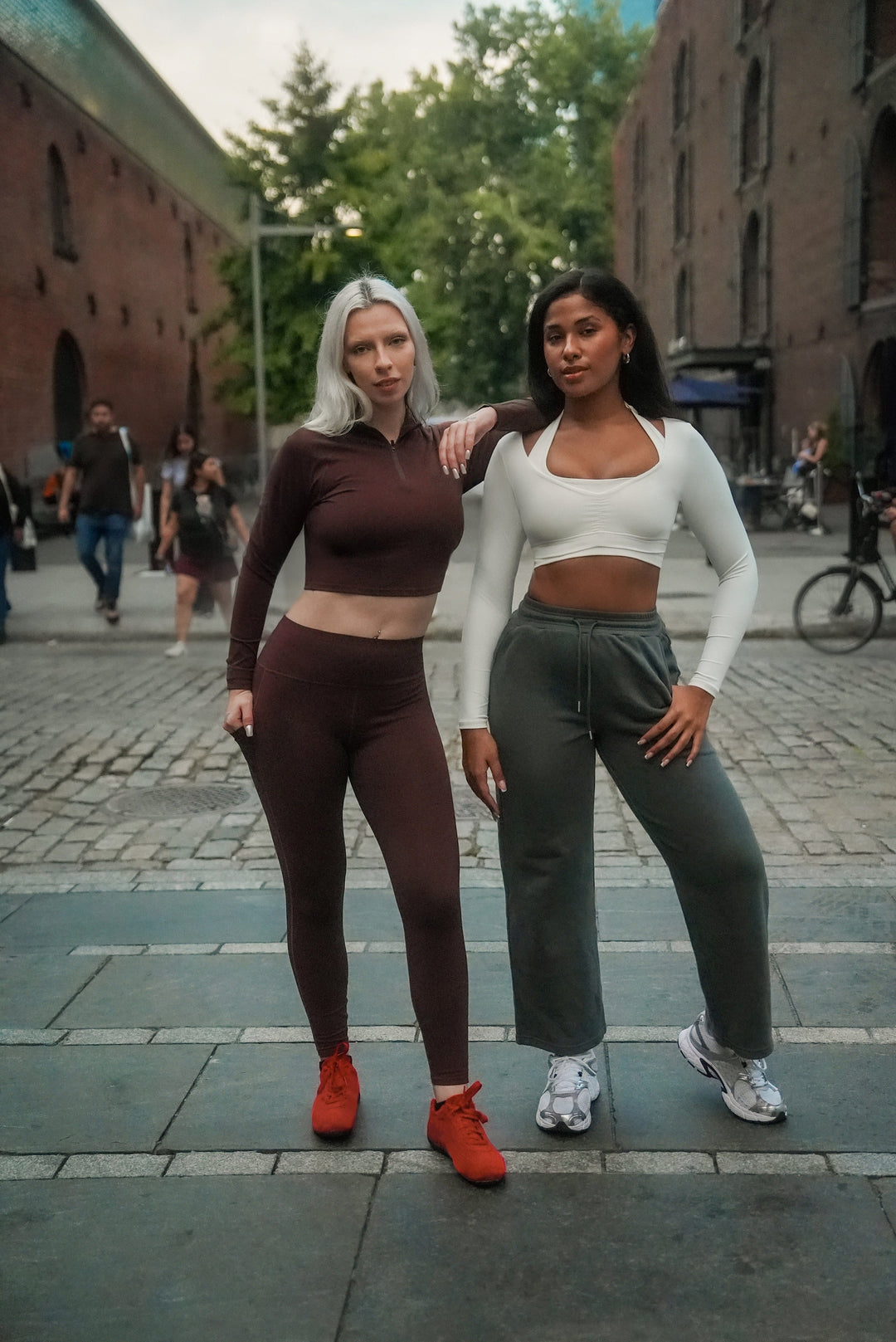 Two women posing on a street with brick buildings and trees in the background